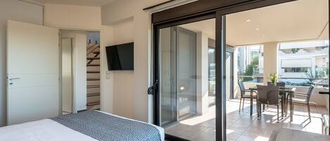 A modern hotel room with a bed, a TV mounted on the wall, a sliding glass door leading to a balcony with outdoor seating.