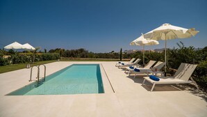 Une piscine entourée de verdure, avec des chaises longues et des parasols, sous un ciel dégagé.