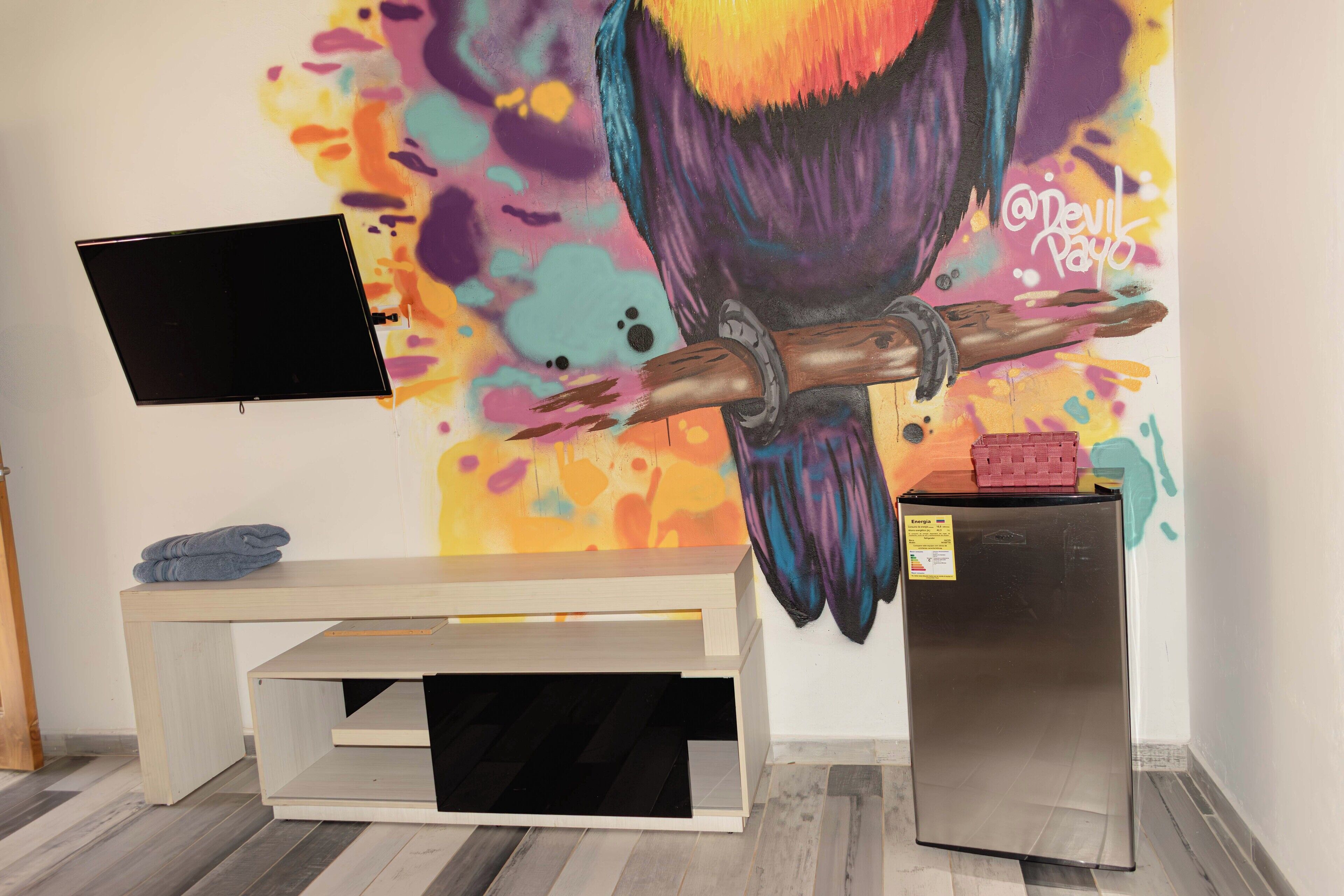 A room with a colorful mural, a flat-screen TV, a wooden bench, and a small refrigerator.