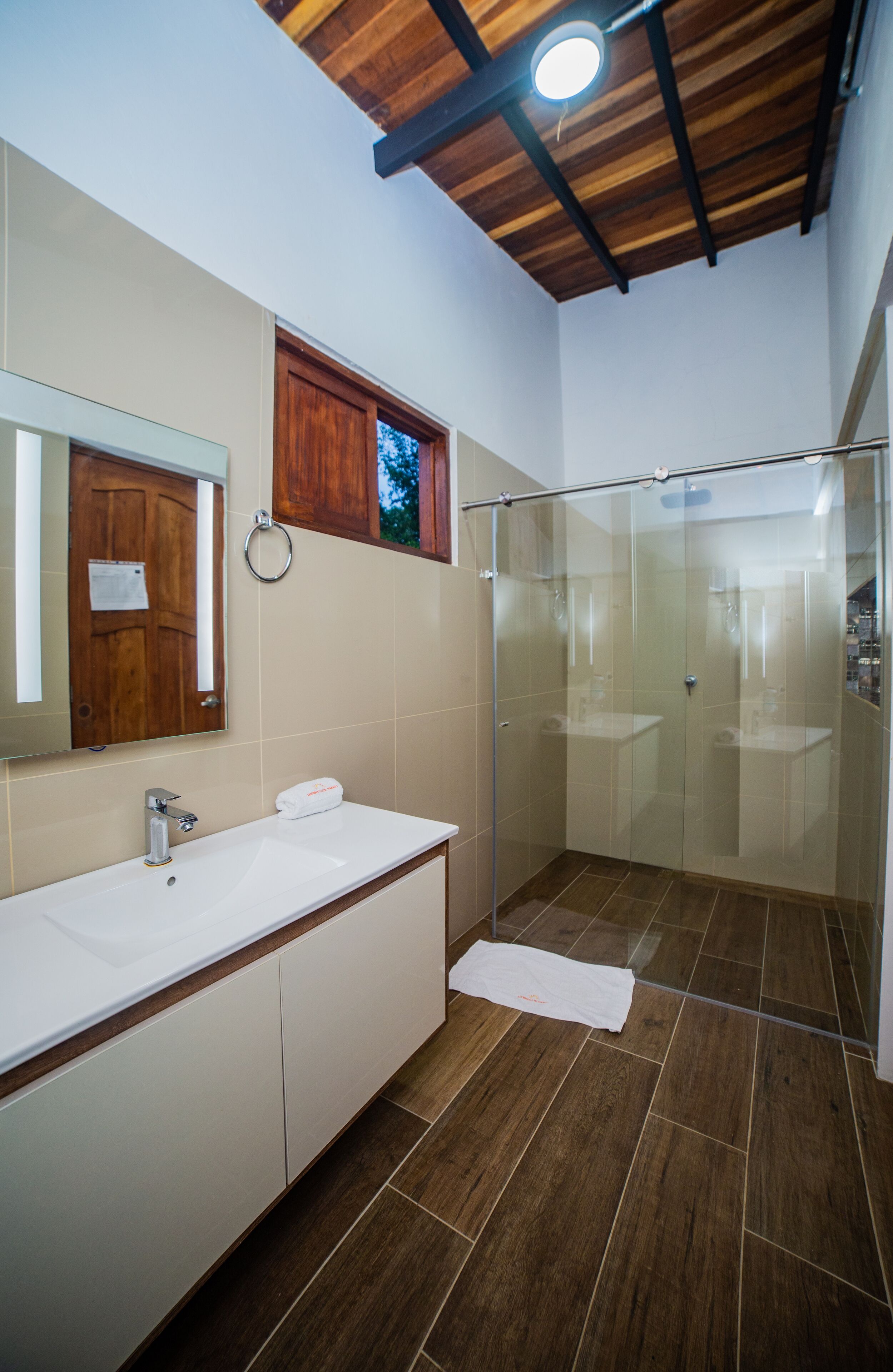 A modern bathroom with a wooden ceiling, a glass shower enclosure, a white sink, and a mirror.
