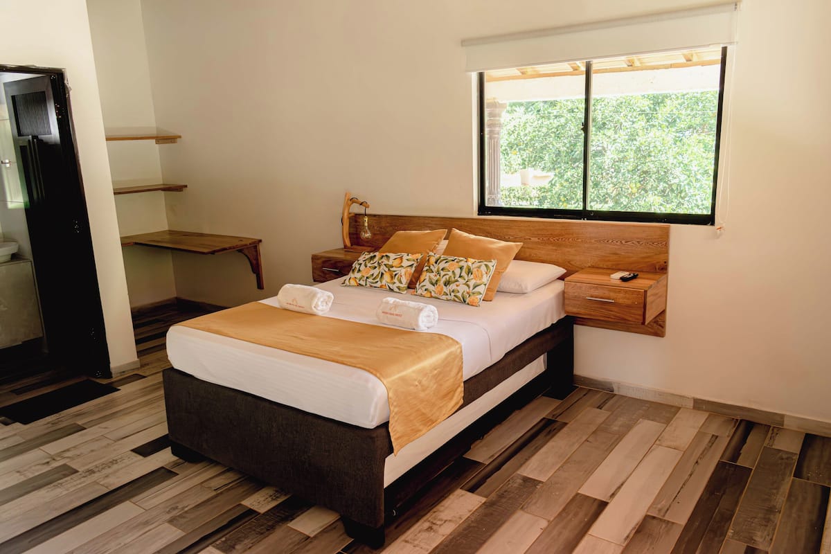 Una cama bien hecha con cabecera de madera, una mesita de noche y una ventana con vistas a la vegetación.