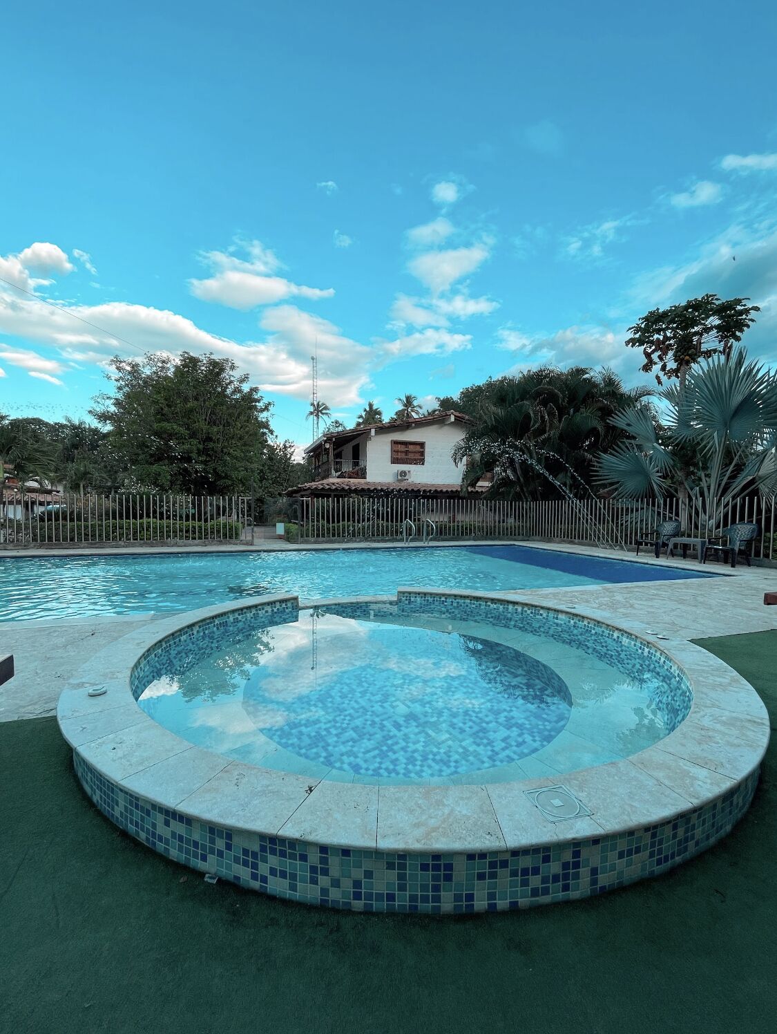 A swimming pool with a circular hot tub area, surrounded by a fence and palm trees.