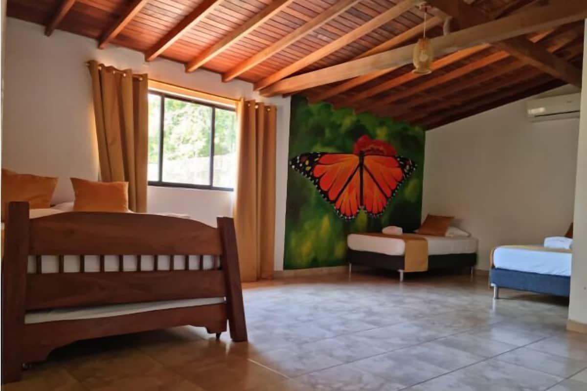 Un dormitorio con techo de madera, una cama con cabecera, una ventana con cortinas, un mural de una mariposa en la pared y una mesita de noche pequeña.