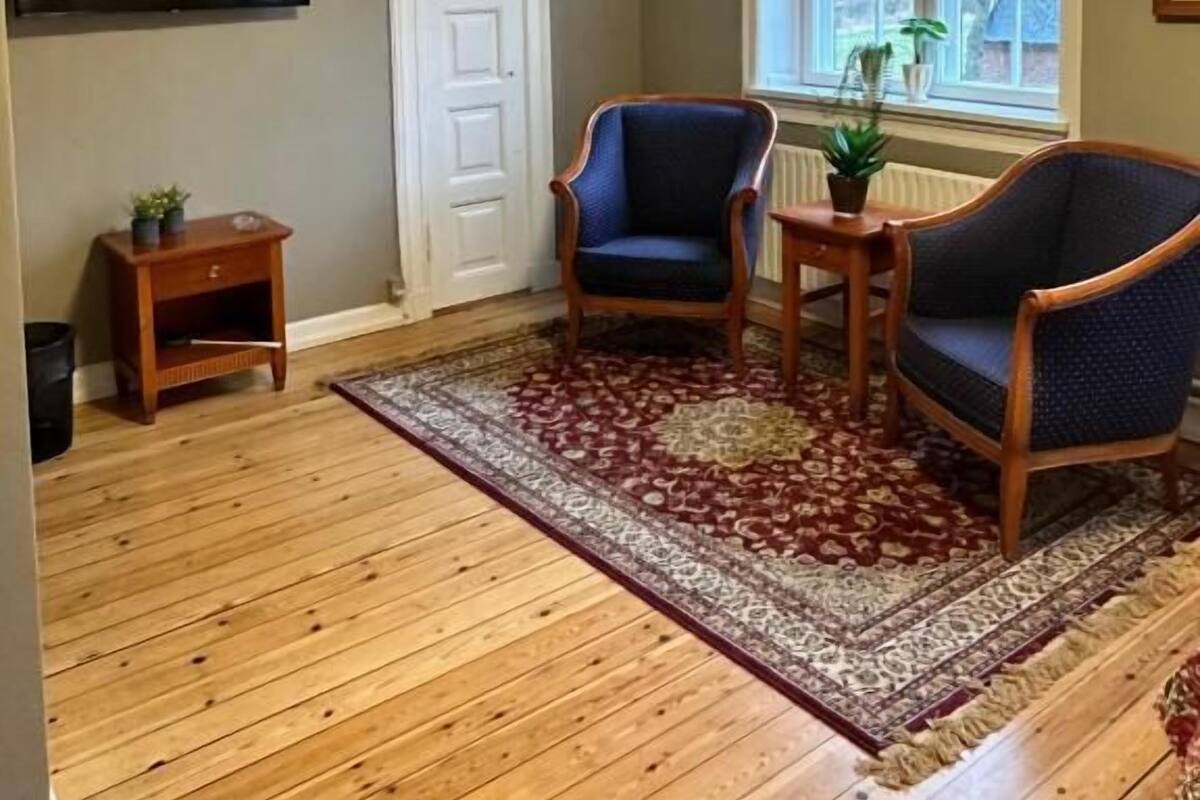 A room with wooden flooring, two blue armchairs, a small wooden table, a TV mounted on the wall, a white door, and a window with a view of trees.
