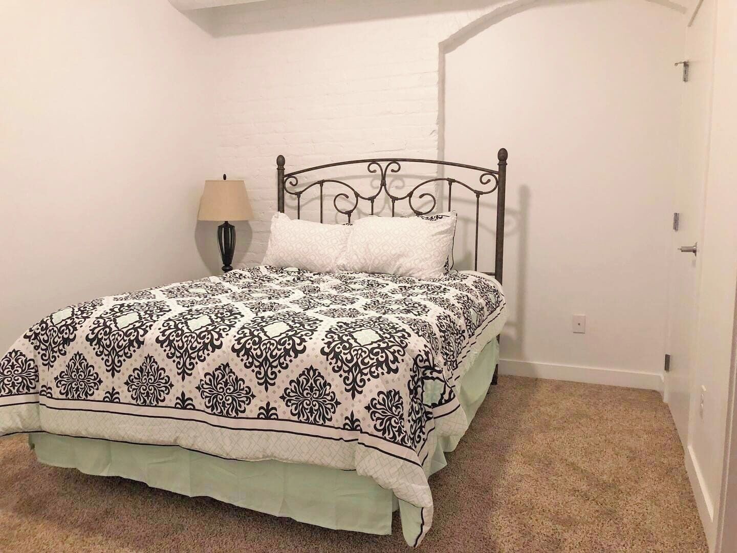 A single bed with a patterned bedspread and two pillows, a bedside lamp, and a white door in a room with a white brick wall and carpeted floor.