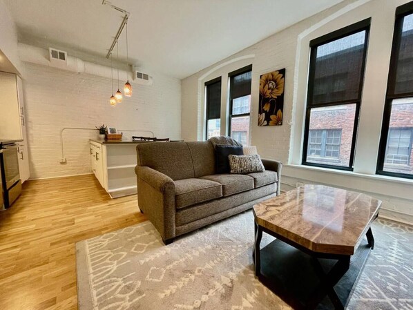A modern living room with a gray sofa, a wooden coffee table, and a patterned rug. There are large windows with black frames and a city view.