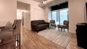 Ein geräumiges Wohnzimmer mit einem dunkelgrauen Sofa, zwei gemusterten Sesseln, Holzboden und einem großen Fenster, das für natürliches Licht sorgt.