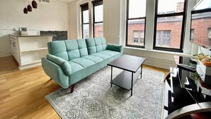 Ein Wohnzimmer mit einem salbeengrünfarbenen Sofa, einem kleinen Couchtisch und einem gemusterten Teppich. Große Fenster bieten Blick auf ein Backsteingebäude.