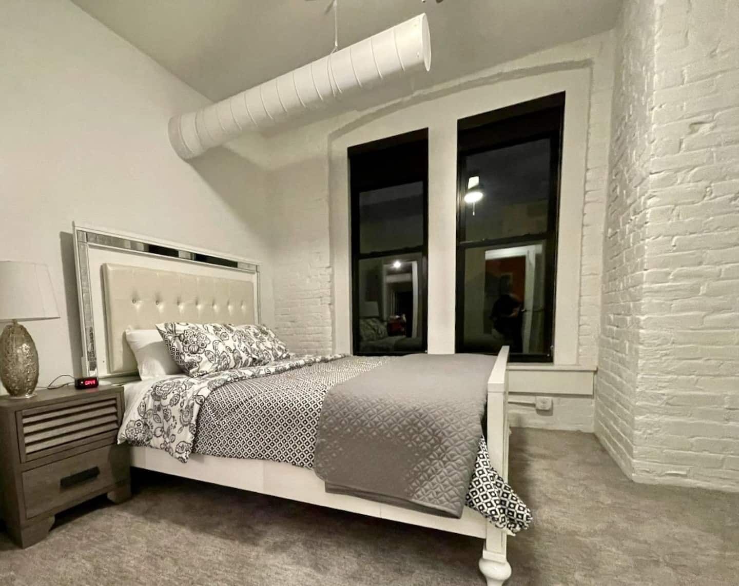 A bedroom with a white bed, a nightstand, a window with a view of the outside, and a white brick wall.