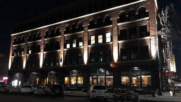 A multi-story building with a brick facade and illuminated windows, featuring a ground-level entrance with a sidewalk and parked cars. - A historic movie themed, 1,200 square foot loft in the heart of the Old Market (Omaha)