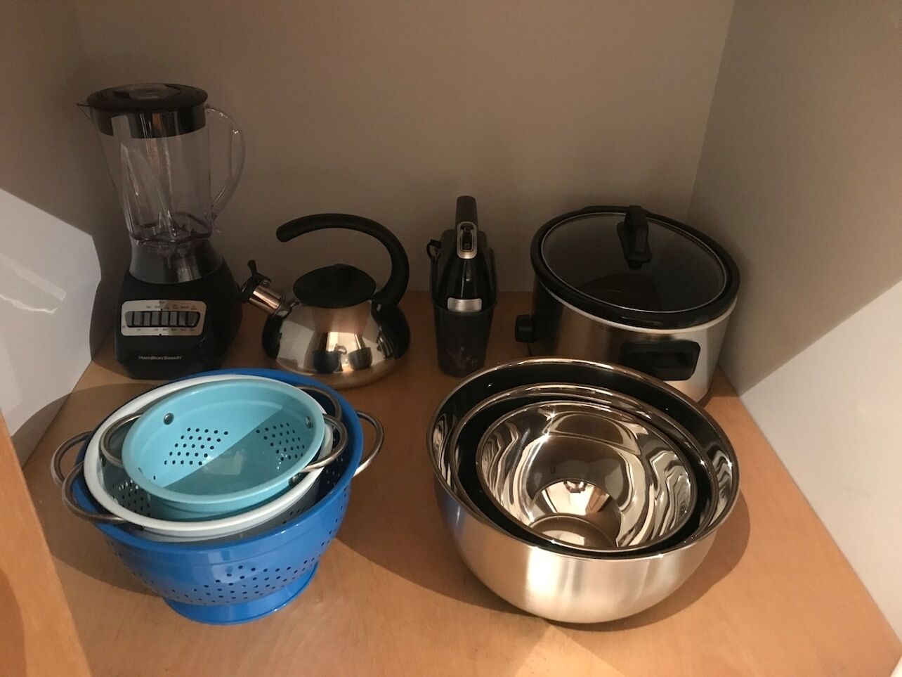 A kitchen counter with a blender, kettle, electric kettle, slow cooker, colander, and stainless steel bowl.