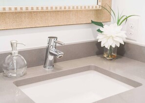 A bathroom sink with a chrome faucet, a clear glass soap dispenser, and a vase with white flowers. - A historic beautifully detailed suite, 1,300 square foot, corner loft in the heart of the Old Market (Omaha)