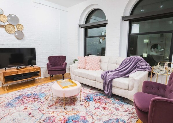 A living room with a white sofa, a purple armchair, a TV on a wooden stand, and a colorful rug. - A historic beautifully detailed suite, 1,300 square foot, corner loft in the heart of the Old Market (Omaha)