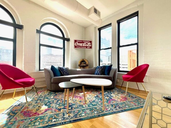Una sala de estar moderna con un sofá, dos sillas rojas, una mesa de centro y una alfombra colorida. Hay ventanas amplias y un cartel de Coca-Cola en la pared.
