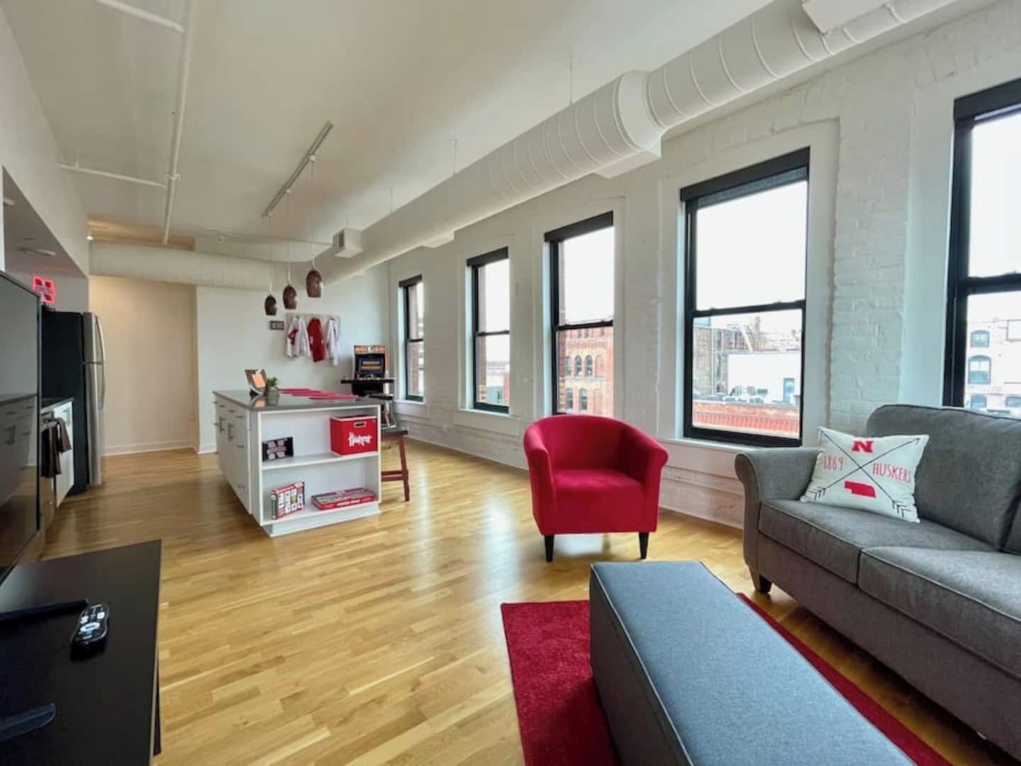 Un appartement moderne doté d’une cuisine, d’un fauteuil rouge, d’un canapé gris et de grandes fenêtres offrant une vue sur la ville.