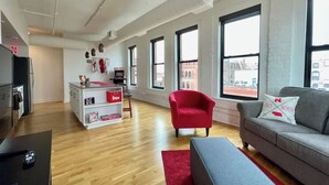 Un apartamento moderno con cocina, un sillón rojo, un sofá gris y ventanas amplias que ofrecen vistas a la ciudad.