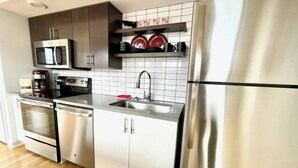 Una cocina moderna con electrodomésticos de acero inoxidable, microondas, horno y lavavajillas. Hay un fregadero con grifo, y una repisa encima con platos y tazas.