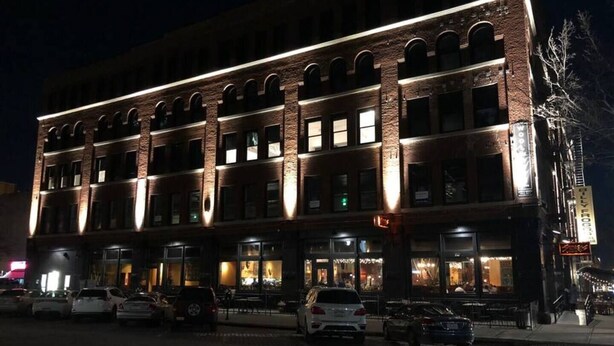 Un edificio de varios pisos con fachada de ladrillo y ventanas iluminadas, que cuenta con un restaurante en la planta baja y mesas al aire libre.