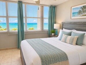 A bedroom with a large bed, a nightstand with a lamp, a framed picture on the wall, and a view of the ocean through the window.