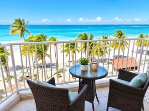 A balcony with wicker furniture and a small table, overlooking a beach with palm trees and the ocean.