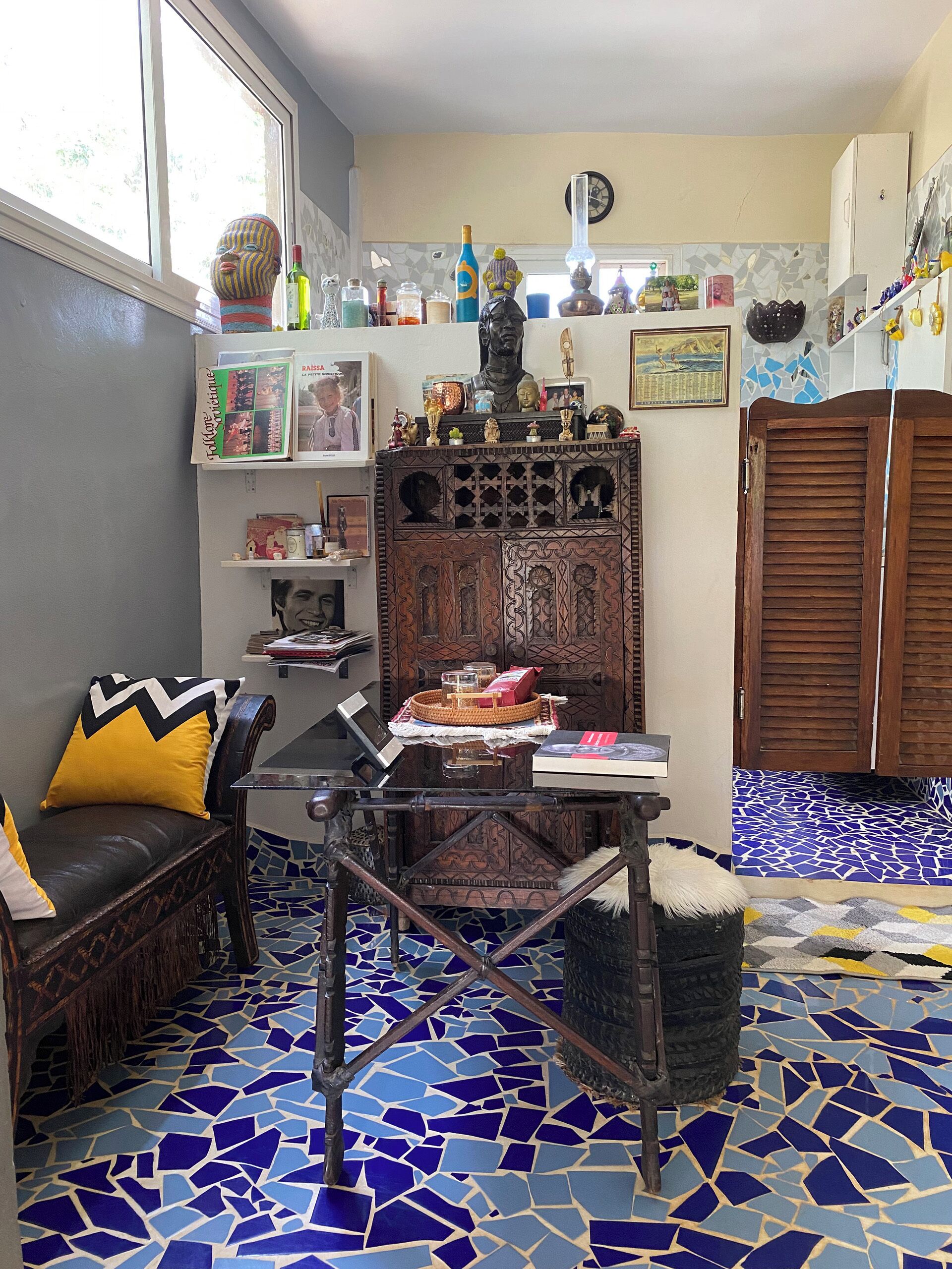 Habitació amb terra de mosaic blau, un armari de fusta amb objectes decoratius, un sofà amb coixins grocs i una taula petita amb un ordinador portàtil i llibres.