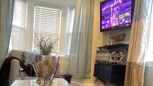 A living room with a glass-top coffee table, a sofa, a TV mounted on the wall, and a window with blinds. - Charming 4 Br 2Ba Duplex  (Baltimore)