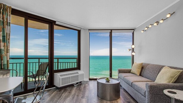 A balcony with a view of the ocean, a gray sofa, a small round table, and a chair. - Sleek Oceanfront King Suite! Dogs OK (Myrtle Beach)
