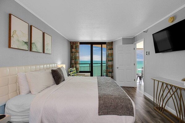 A hotel room with a large bed, a TV mounted on the wall, and a balcony with a view of the ocean.