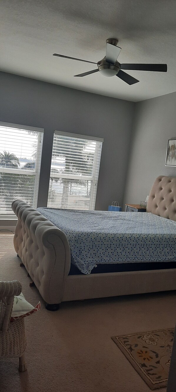 A bedroom with a bed, a ceiling fan, a window with blinds, and a view of palm trees outside.