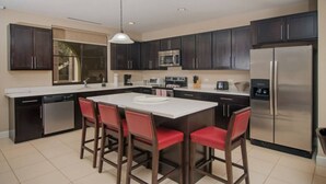 Une cuisine moderne avec des meubles de couleur sombre, un réfrigérateur en acier inoxydable, un four à micro-ondes et un grand îlot central doté de tabourets de bar rouges.