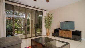 Un salon moderne comprenant un canapé, une table basse en verre, un téléviseur à écran plat posé sur un meuble en bois, et une porte-fenêtre coulissante donnant sur un balcon avec vue sur la verdure.