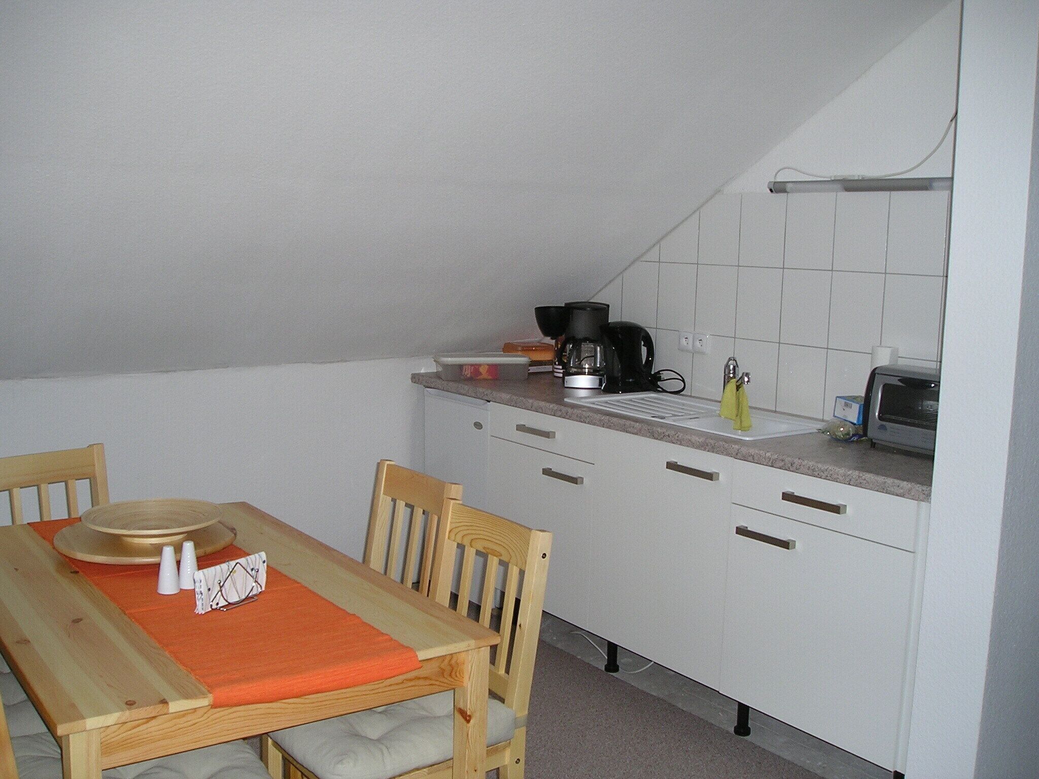 A small kitchen with white cabinets, a wooden dining table with orange placemats, and basic appliances like a coffee maker and toaster.
