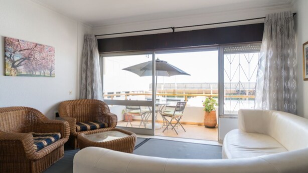 Een kamer met een witte bank, twee rieten stoelen, een klein tafeltje en een balkon met een tafel en stoelen.