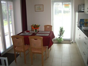 A dining area with a table set for four, chairs, a vase with flowers, and a view of the outside through a window with blinds.