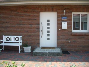 A brick building with a white door, a bench, a potted plant, and a window with blinds.