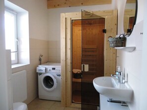 Un baño pequeño con lavadora, una puerta de cristal que da a una sauna y un lavabo blanco.