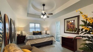 Une chambre à coucher comprenant un lit, des tables de chevet, un ventilateur de plafond, une fenêtre, une porte, un bureau, une chaise et une plante.