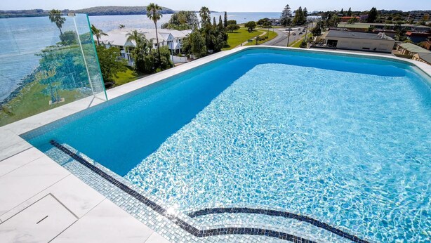 A rectangular swimming pool with clear blue water, surrounded by a tiled deck and a glass barrier. - Oceanside Apartment with Bay Views (Batemans Bay)
