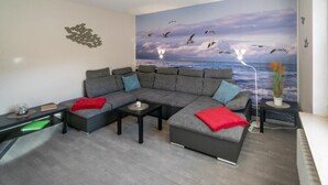 A modern living room with a sectional sofa, a coffee table, and a mural of birds flying over the ocean. - Vacation apartment Möwenblick (Norden)