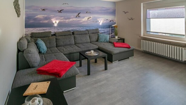 A living room with a grey sectional sofa, a small table with a tray, and a red throw blanket. - Vacation apartment Möwenblick (Norden)