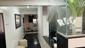A hotel reception area with a glass partition, a reception desk, a vending machine, and a small seating area with patterned ottomans. - HOTEL CENTRAL BARAO DO RIO BRANCO  (Anapolis)