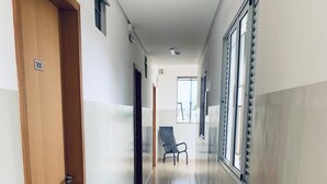 A hallway with a wooden door on the left, a chair in the center, and windows on the right. - HOTEL CENTRAL BARAO DO RIO BRANCO  (Anapolis)