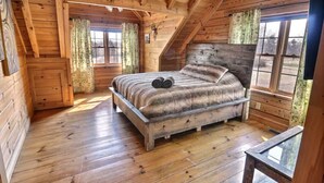 Quarto de cabana de madeira com cama, duas janelas com cortinas e teto de madeira.