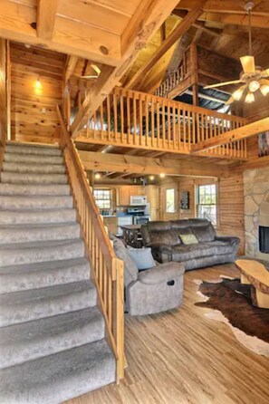 A spacious living room with a stone fireplace, a large sofa, a wooden staircase, and a ceiling fan. - Rustic Cabin Near Purdue University (West Lafayette)