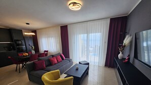 A modern living room with a sofa, a yellow armchair, a coffee table, and a TV on a stand. There is a dining area with a table and chairs, and a kitchen area with a stove and oven. - APARTMENTS GROZIC **** OPATIJA - APARTMENT A2 (Opatija)