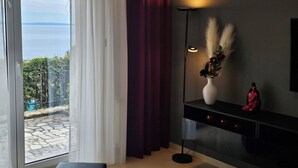 A room with a large sliding glass door leading to a balcony with a view of the sea. There is a floor lamp with a vase of flowers on a black console table. - APARTMENTS GROZIC **** OPATIJA - APARTMENT A2 (Opatija)