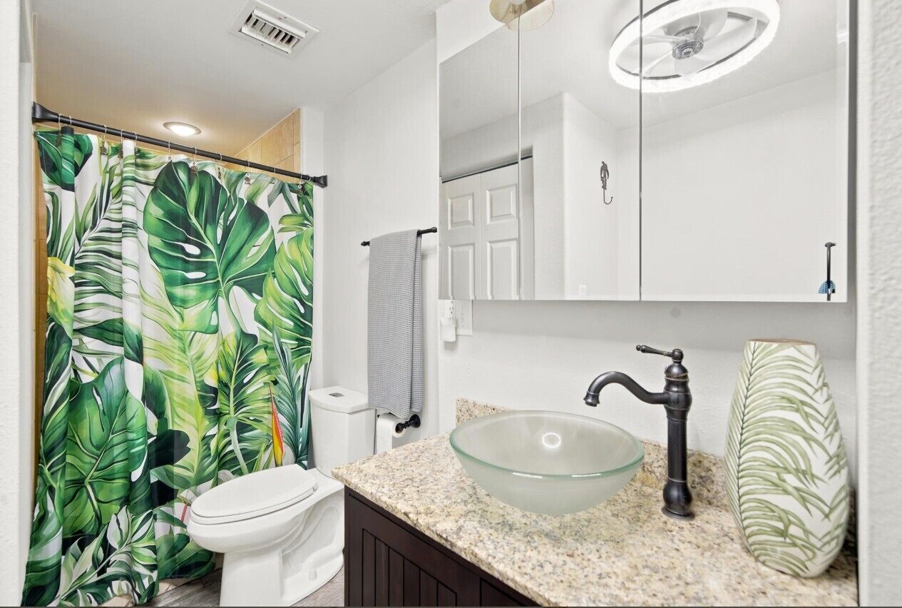 A bathroom with a toilet, a glass vessel sink, a mirror, and a shower curtain with a tropical leaf pattern.