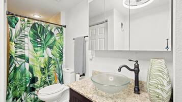 A bathroom with a toilet, a glass vessel sink, a mirror, and a shower curtain with a tropical leaf pattern.