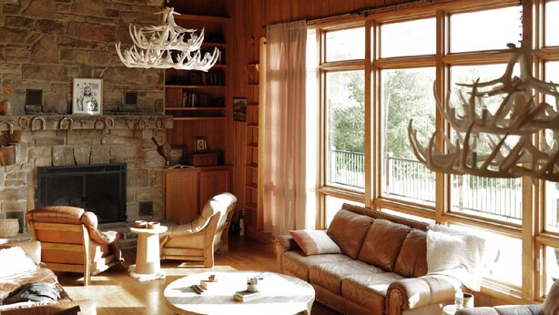 Una sala de estar acogedora con chimenea de piedra, vigas de madera, un candelabro, sofás de cuero y un ventanal con vistas.
