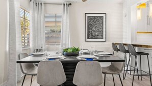 A modern dining area with a rectangular table set for four, grey chairs, a potted plant, and a framed abstract artwork on the wall.